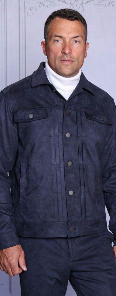 A man wearing the INSOMNIA ROMEO 2PC Vegan PU Suede Casual Jacket & Pants Navy Set by Insomnia over a white turtleneck stands indoors against a light-colored background.