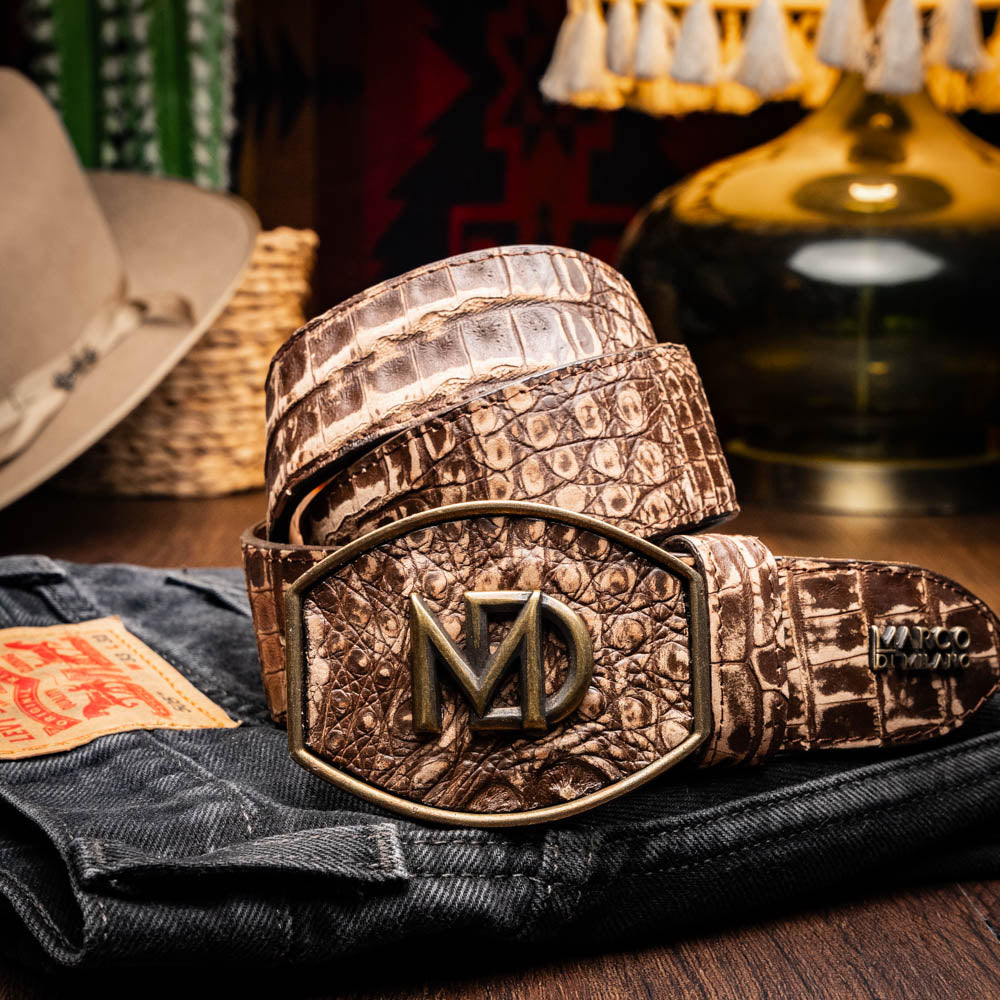 The Marco Di Milano Iowa Belt Stone Brown features genuine caiman crocodile leather, a gold "MD" buckle, and "Marco Milano" engraved on the side, evoking a classic western style against a white background.