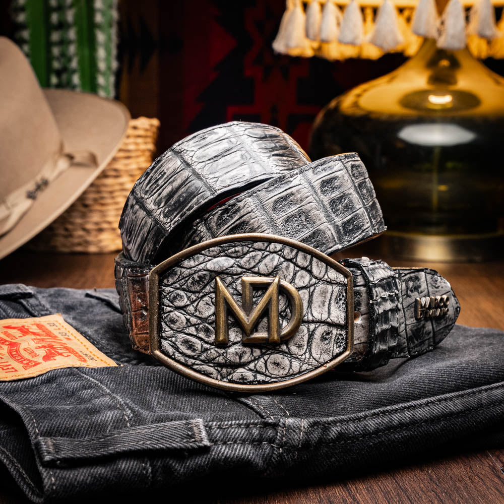 The Marco Di Milano Iowa Belt Stone Gray features genuine caiman skin and a bold gold "MD" buckle on a textured background, combining luxury with standout Western style.