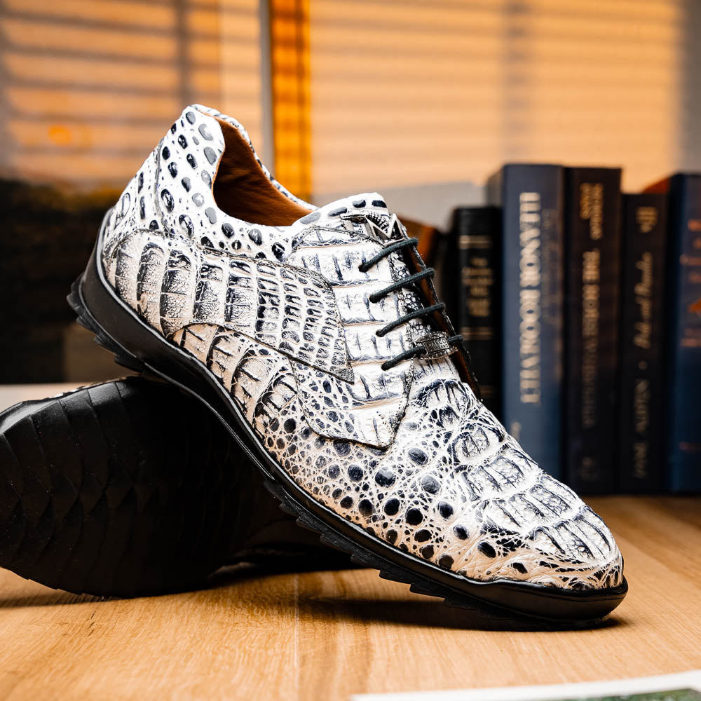 The Marco Di Milano Kratos Newspaper features white and black crocodile-patterned luxury dress shoes with black soles; one shoe stands upright, while the other lies on its side showcasing the hand-painted finish.