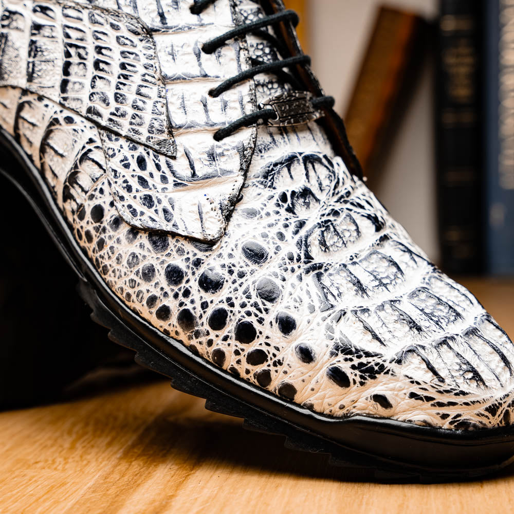 The Marco Di Milano Kratos Newspaper features white and black crocodile-patterned luxury dress shoes with black soles; one shoe stands upright, while the other lies on its side showcasing the hand-painted finish.