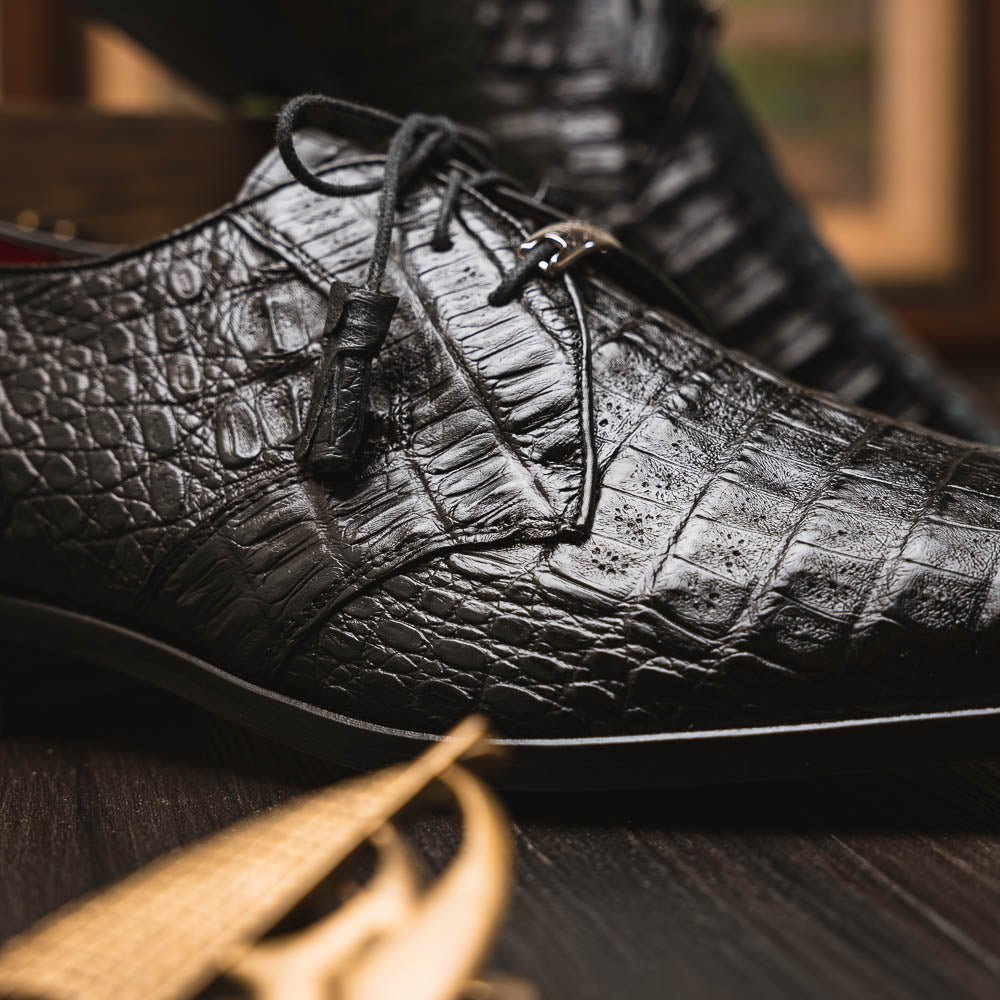 A pair of Marco Di Milano Lacio Black shoes—crafted from genuine caiman crocodile leather—one upright and one on its side, showing a brown leather sole embossed with luxury branding.