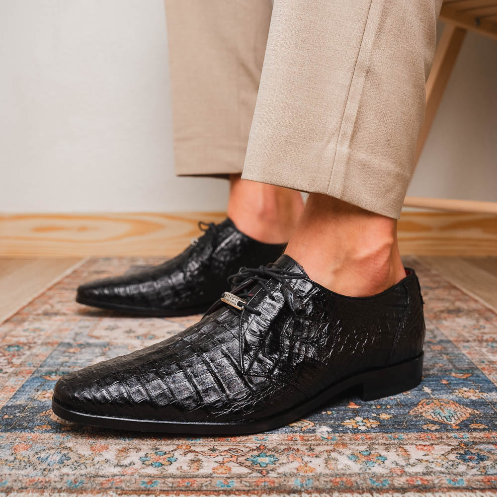 A pair of Marco Di Milano Lacio Black shoes—crafted from genuine caiman crocodile leather—one upright and one on its side, showing a brown leather sole embossed with luxury branding.
