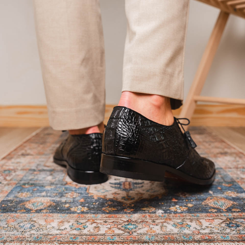 A pair of Marco Di Milano Lacio Black shoes—crafted from genuine caiman crocodile leather—one upright and one on its side, showing a brown leather sole embossed with luxury branding.