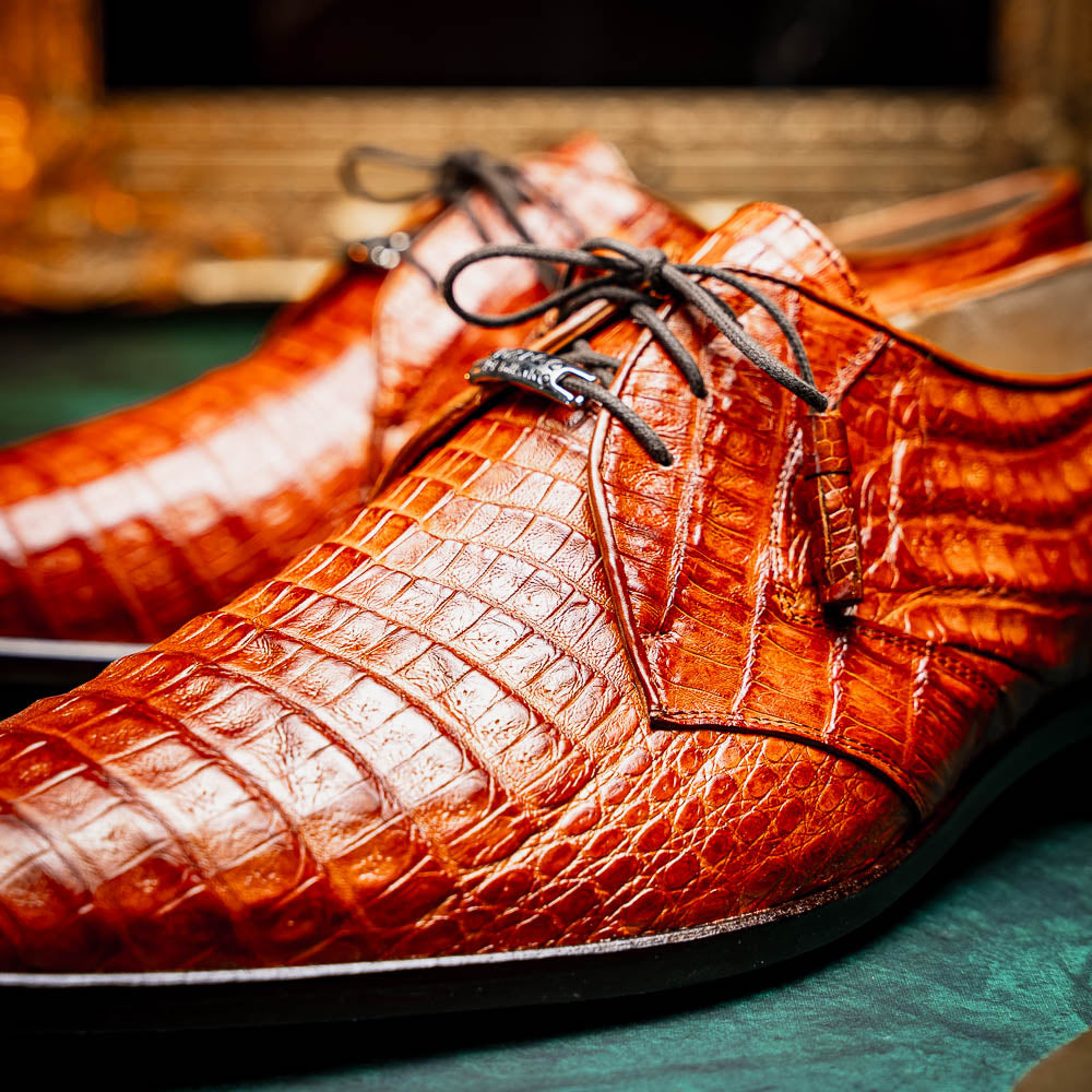 A pair of Marco Di Milano Lacio Brandy luxury derby shoes in a brown crocodile pattern, crafted from genuine caiman leather—one upright and one on its side to highlight the signature sole branding.