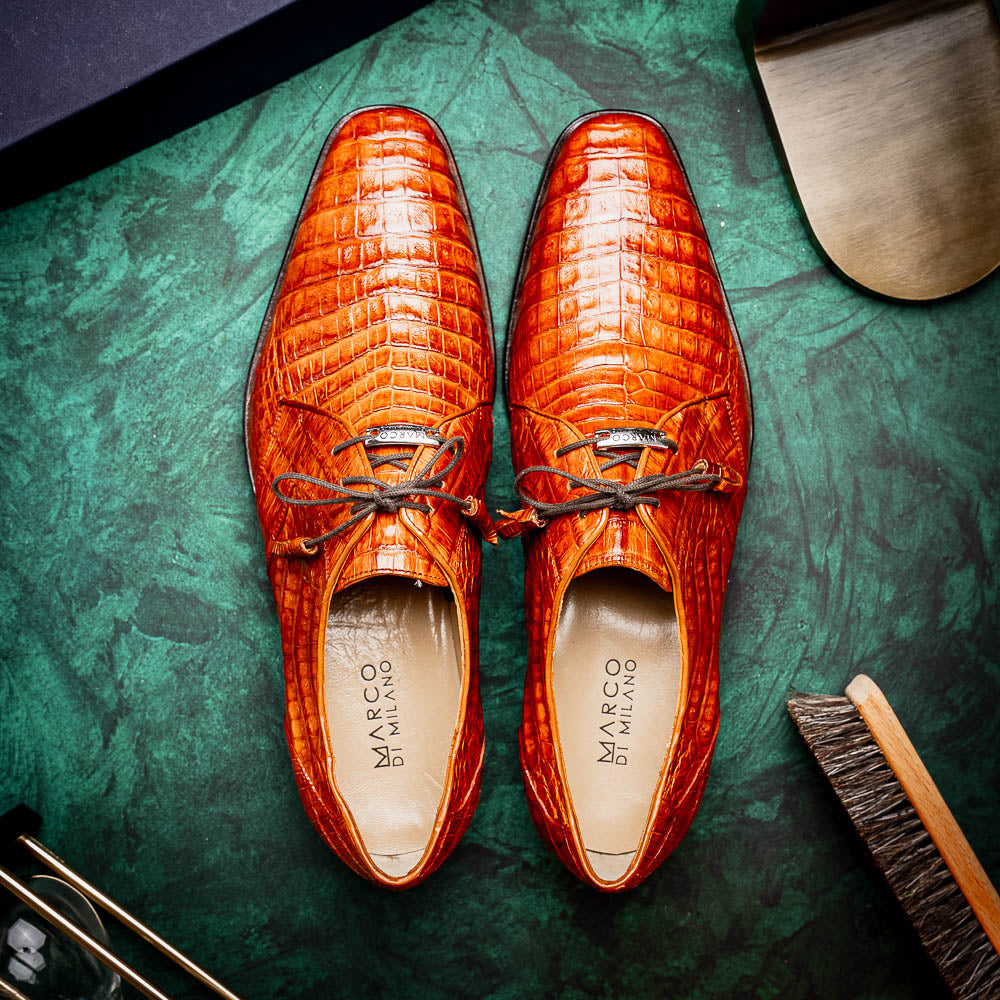 A pair of Marco Di Milano Lacio Brandy luxury derby shoes in a brown crocodile pattern, crafted from genuine caiman leather—one upright and one on its side to highlight the signature sole branding.
