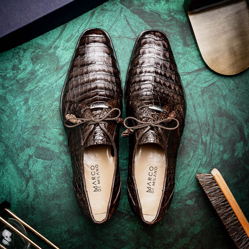 The "Lacio Brown" by Marco Di Milano is a luxury men's dress shoe made from genuine caiman crocodile leather, featuring tassel laces and the brand's logo—presented with one shoe resting atop the other.