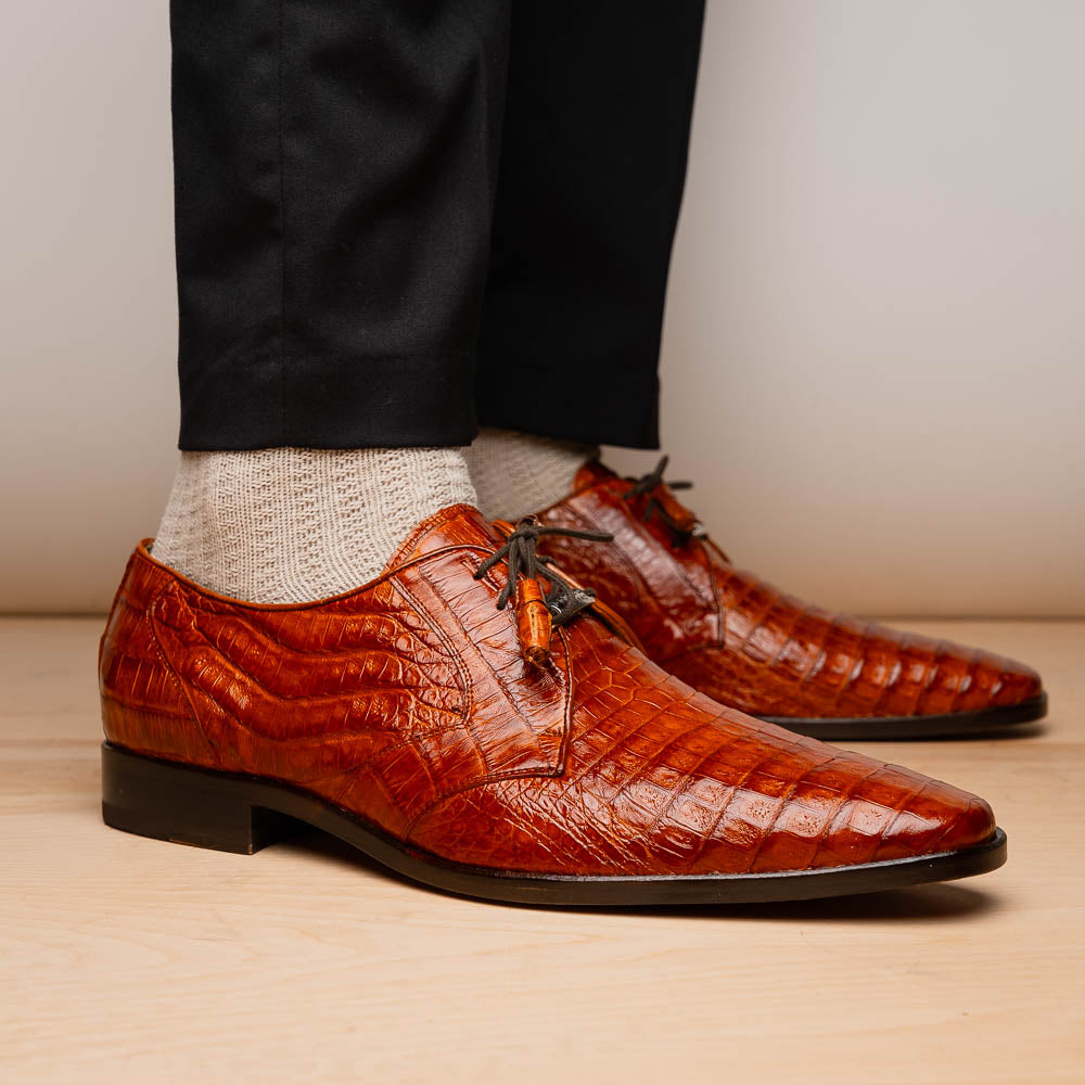 A pair of Marco Di Milano Lacio Brandy luxury derby shoes in a brown crocodile pattern, crafted from genuine caiman leather—one upright and one on its side to highlight the signature sole branding.
