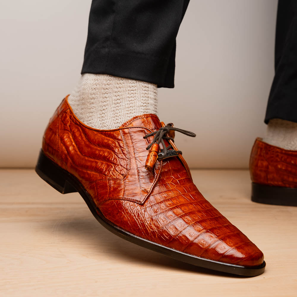 A pair of Marco Di Milano Lacio Brandy luxury derby shoes in a brown crocodile pattern, crafted from genuine caiman leather—one upright and one on its side to highlight the signature sole branding.