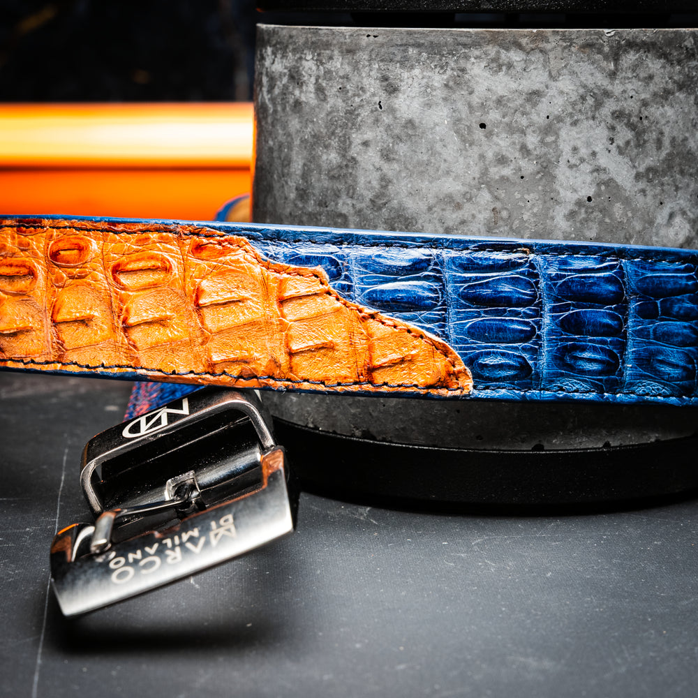 The Luciano Belt in Blue and Brandy by Marco Di Milano—both crafted from genuine caiman leather and adorned with silver buckles—are elegantly coiled together against a white background.