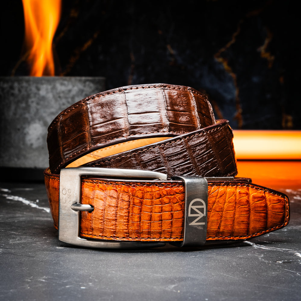 Two handcrafted men’s belts from Marco Di Milano—the Luciano Belt in Brown and Cognac—are coiled and stacked, each highlighted by a silver buckle. These premium accessories showcase the refined elegance and distinctive character of genuine caiman leather, perfect for those who value superior quality.