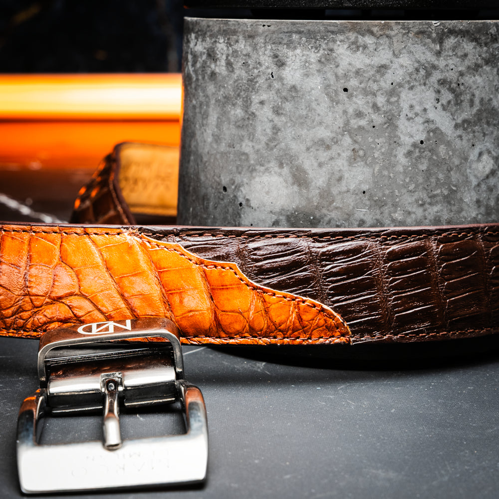 Two handcrafted men’s belts from Marco Di Milano—the Luciano Belt in Brown and Cognac—are coiled and stacked, each highlighted by a silver buckle. These premium accessories showcase the refined elegance and distinctive character of genuine caiman leather, perfect for those who value superior quality.