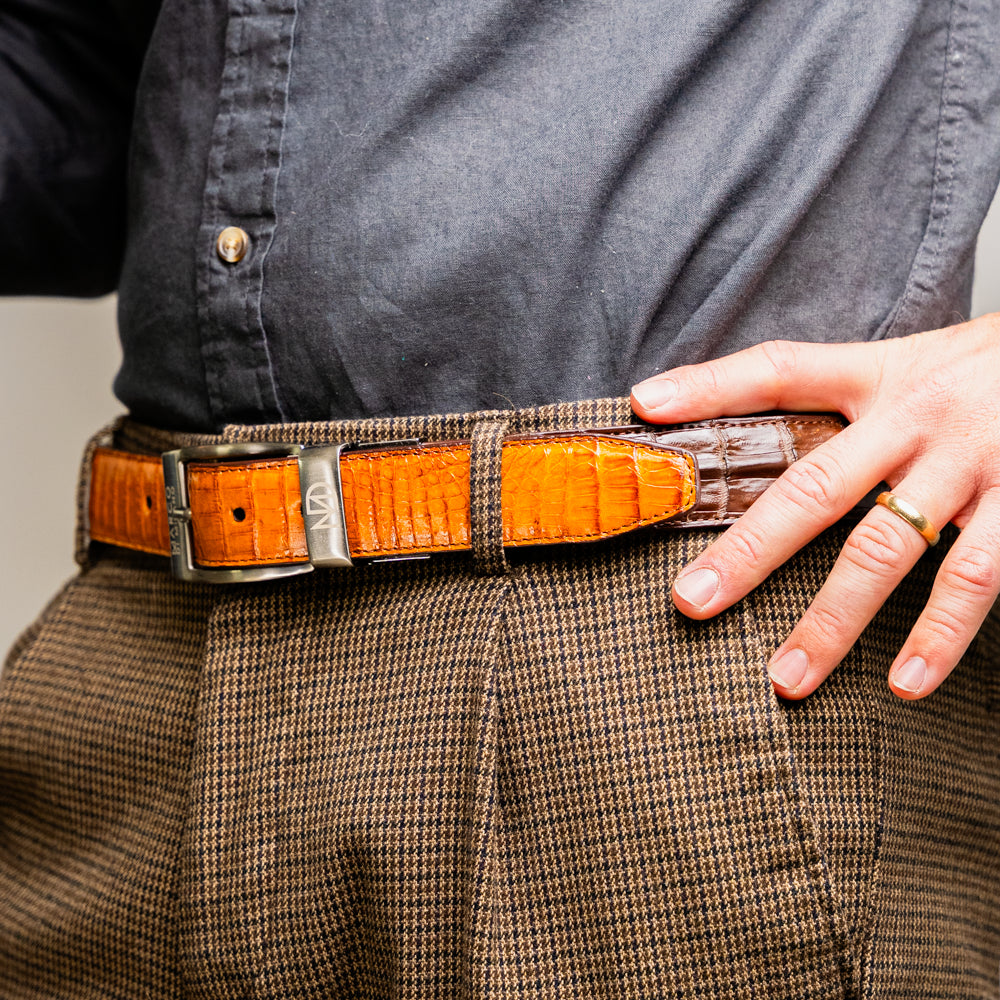 Two handcrafted men’s belts from Marco Di Milano—the Luciano Belt in Brown and Cognac—are coiled and stacked, each highlighted by a silver buckle. These premium accessories showcase the refined elegance and distinctive character of genuine caiman leather, perfect for those who value superior quality.