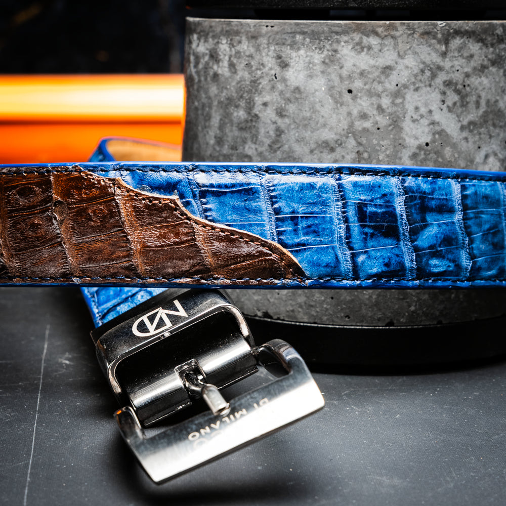 Introducing the Luciano Belt Navy / Brown by Marco Di Milano, a handcrafted luxury belt featuring an exotic crocodile-patterned leather in striking blue and dark brown sections with a refined silver buckle, elegantly coiled on a white background.