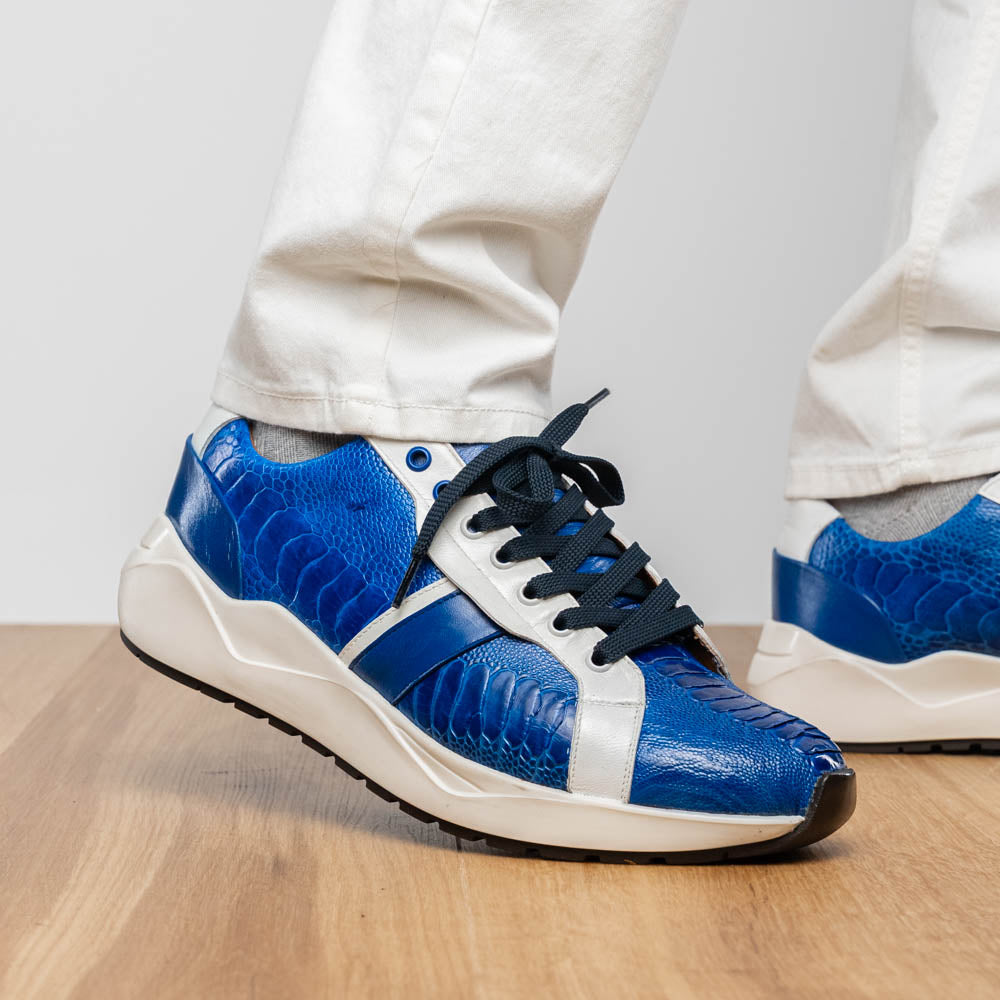 The Marco Di Milano Lyon Blue / White sneakers feature blue and white tones, black laces, textured side panels, and sleek black outsoles, with one shoe displayed stylishly on its side against the other.