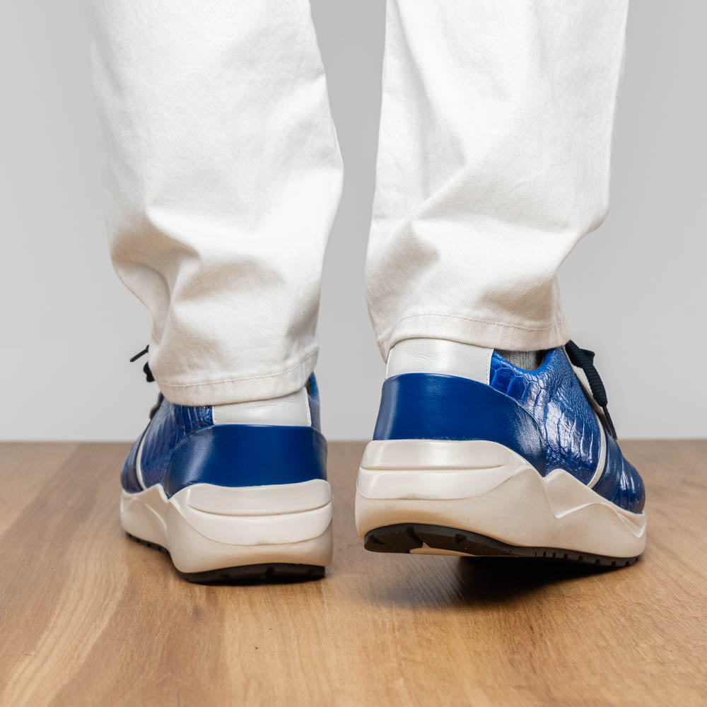 The Marco Di Milano Lyon Blue / White sneakers feature blue and white tones, black laces, textured side panels, and sleek black outsoles, with one shoe displayed stylishly on its side against the other.