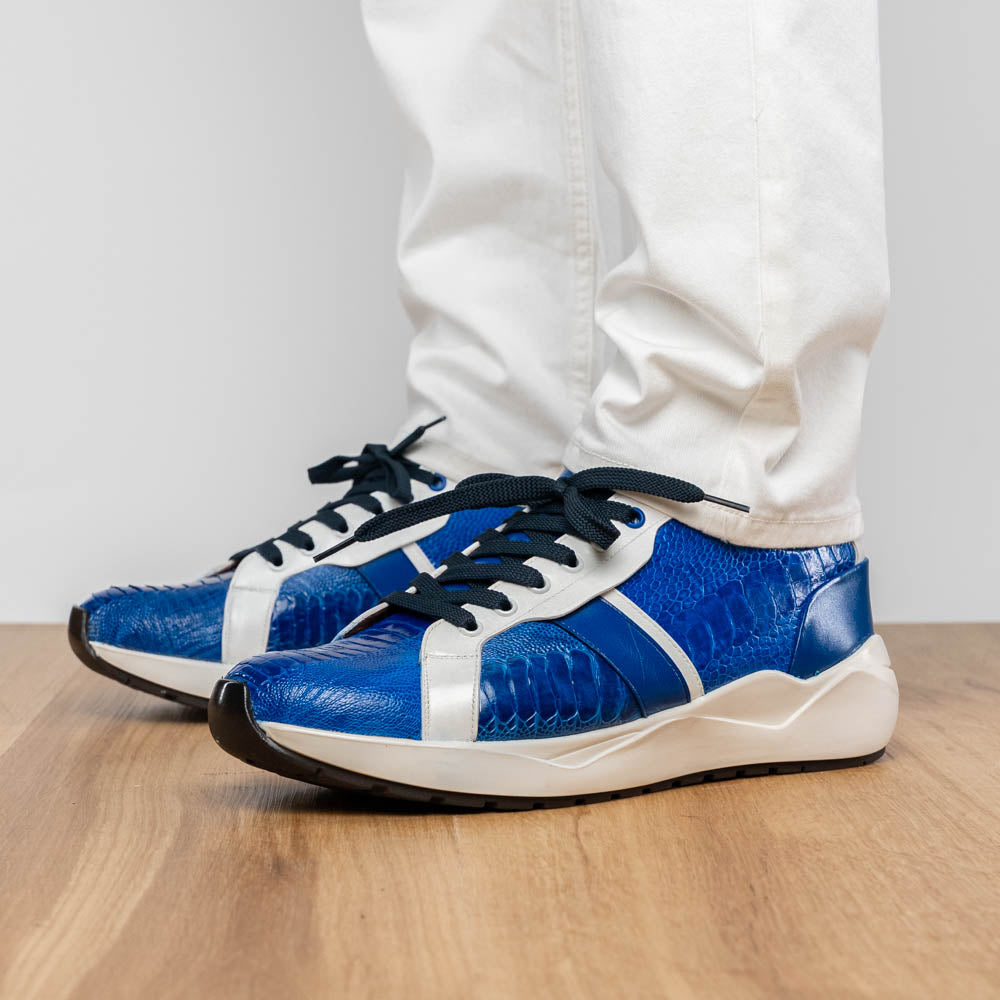 The Marco Di Milano Lyon Blue / White sneakers feature blue and white tones, black laces, textured side panels, and sleek black outsoles, with one shoe displayed stylishly on its side against the other.