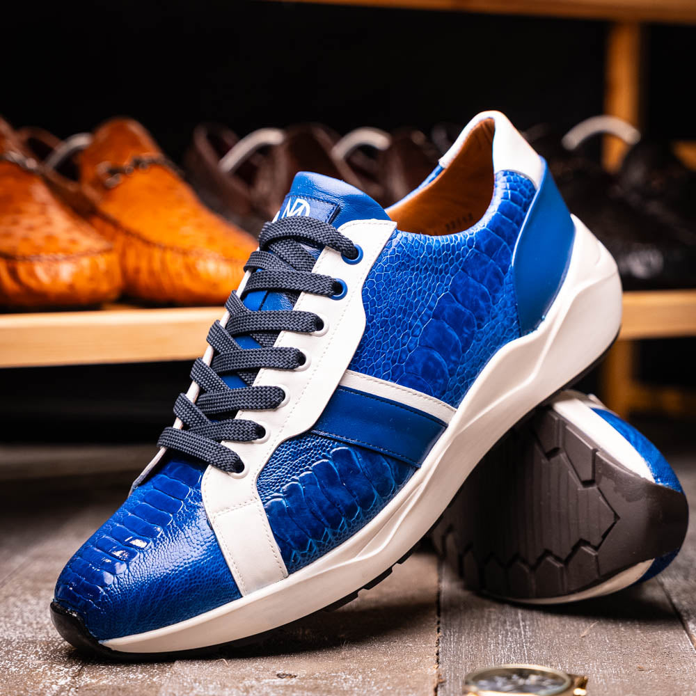 The Marco Di Milano Lyon Blue / White sneakers feature blue and white tones, black laces, textured side panels, and sleek black outsoles, with one shoe displayed stylishly on its side against the other.