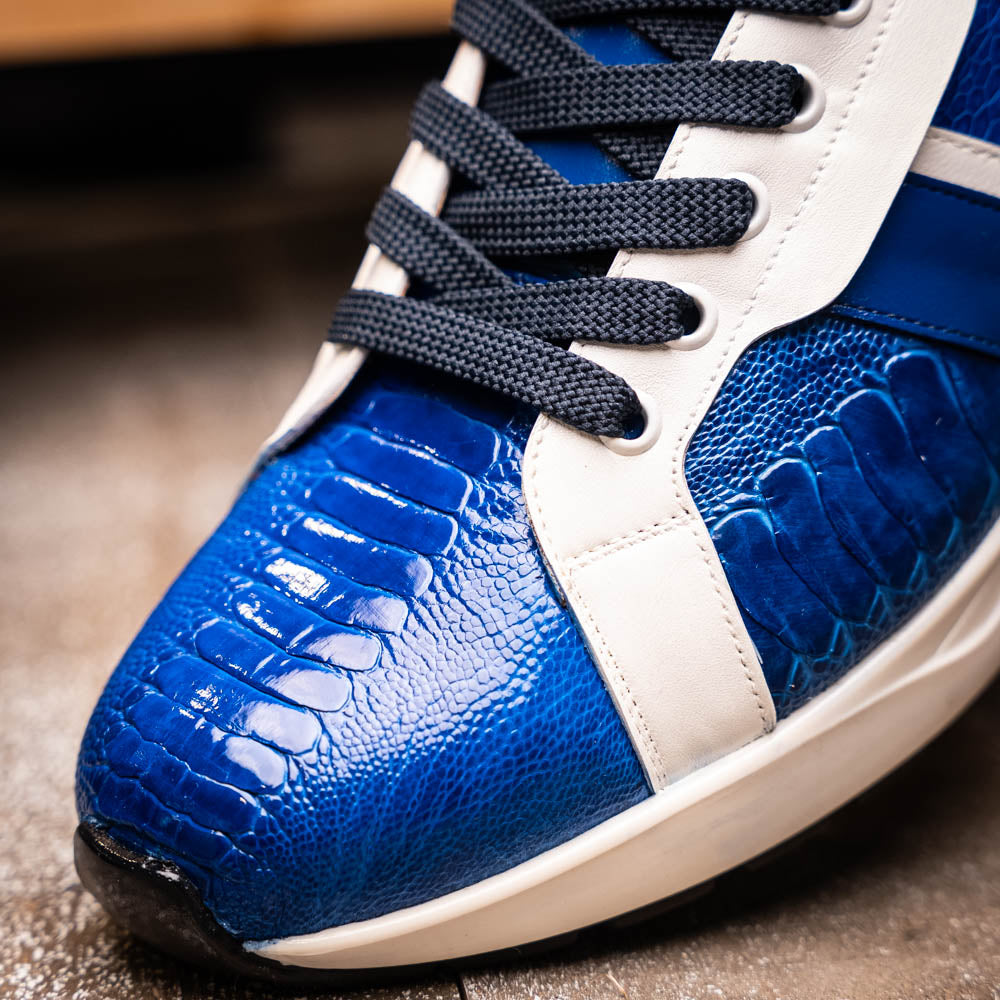 The Marco Di Milano Lyon Blue / White sneakers feature blue and white tones, black laces, textured side panels, and sleek black outsoles, with one shoe displayed stylishly on its side against the other.