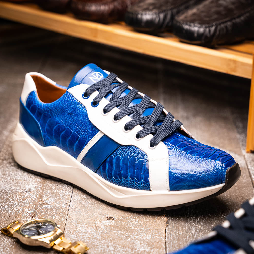 The Marco Di Milano Lyon Blue / White sneakers feature blue and white tones, black laces, textured side panels, and sleek black outsoles, with one shoe displayed stylishly on its side against the other.