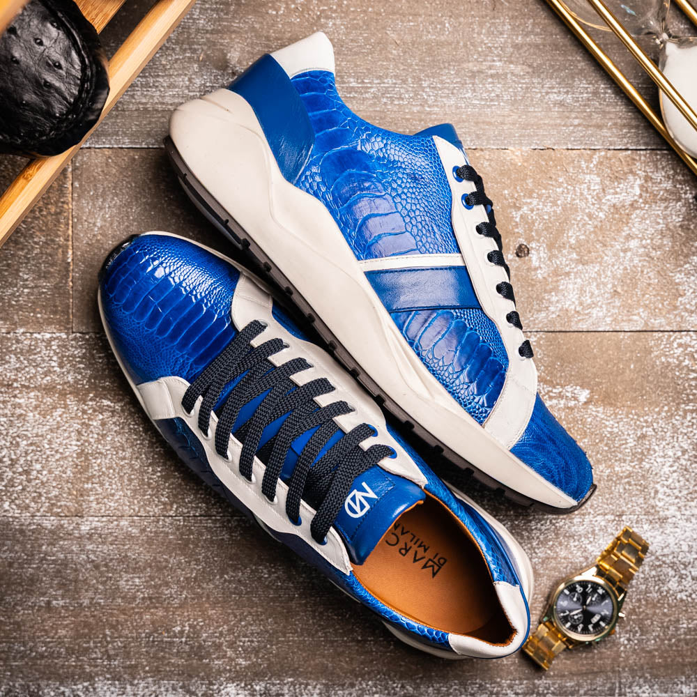 The Marco Di Milano Lyon Blue / White sneakers feature blue and white tones, black laces, textured side panels, and sleek black outsoles, with one shoe displayed stylishly on its side against the other.