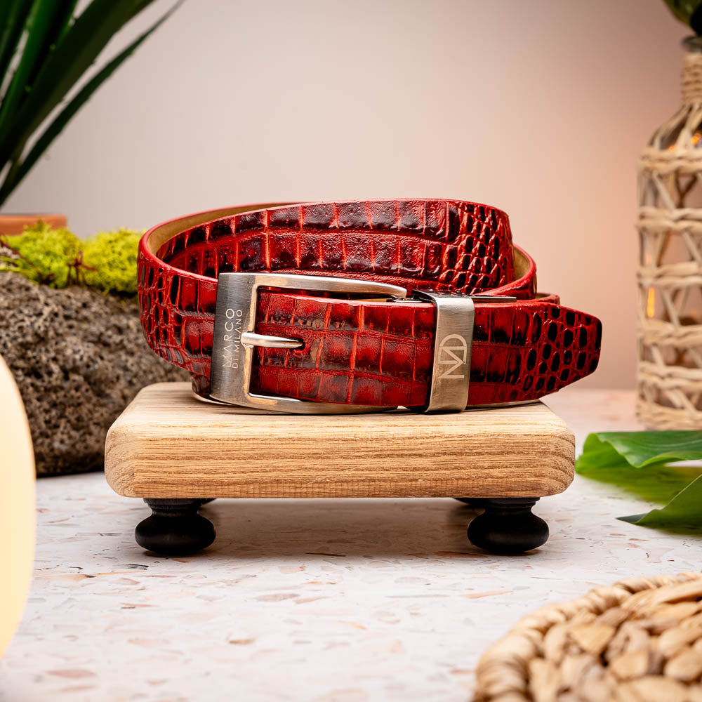 The Emilio Belt Antique Red by Marco Di Milano is a rolled-up red crocodile-patterned leather belt with hand-painted finishes and a silver buckle, displayed on a white background.