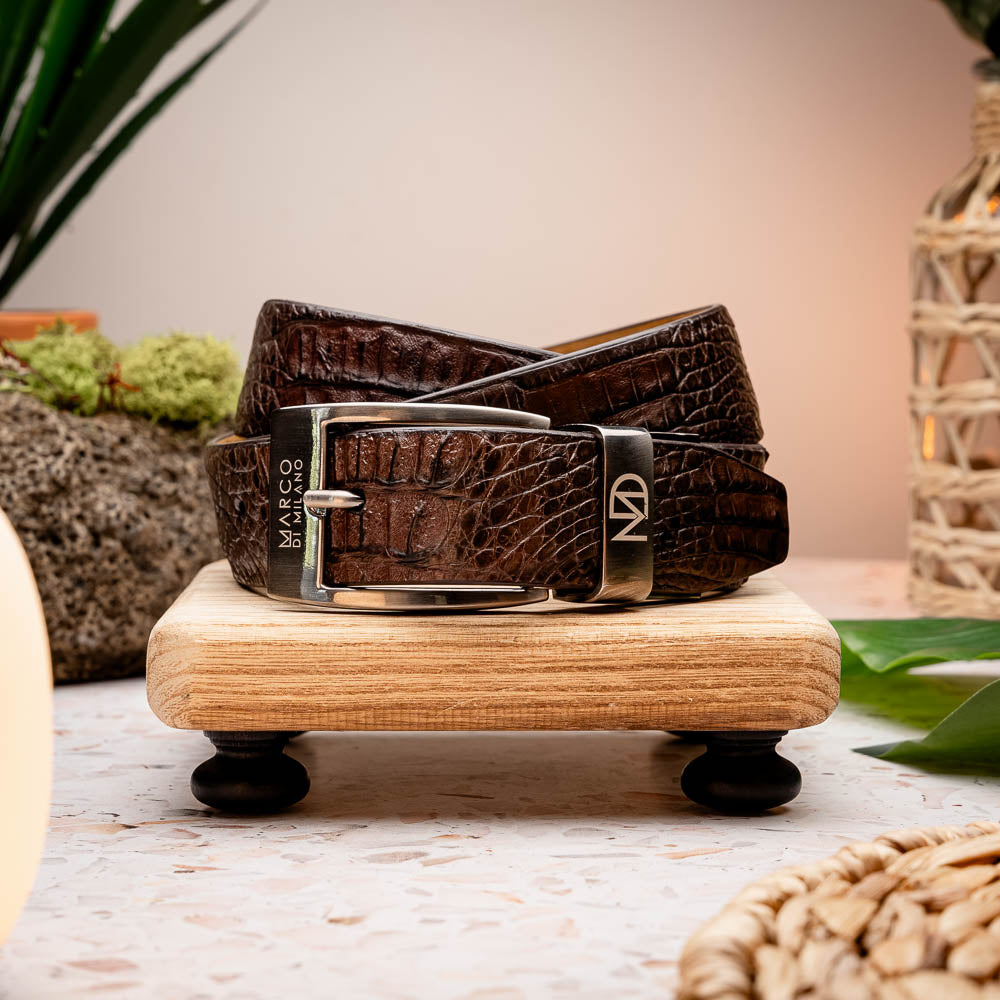 The Riccardo Belt Brown by Marco Di Milano is a dark brown exotic leather belt with a silver buckle and an engraved logo, crafted to mimic genuine caiman belly.