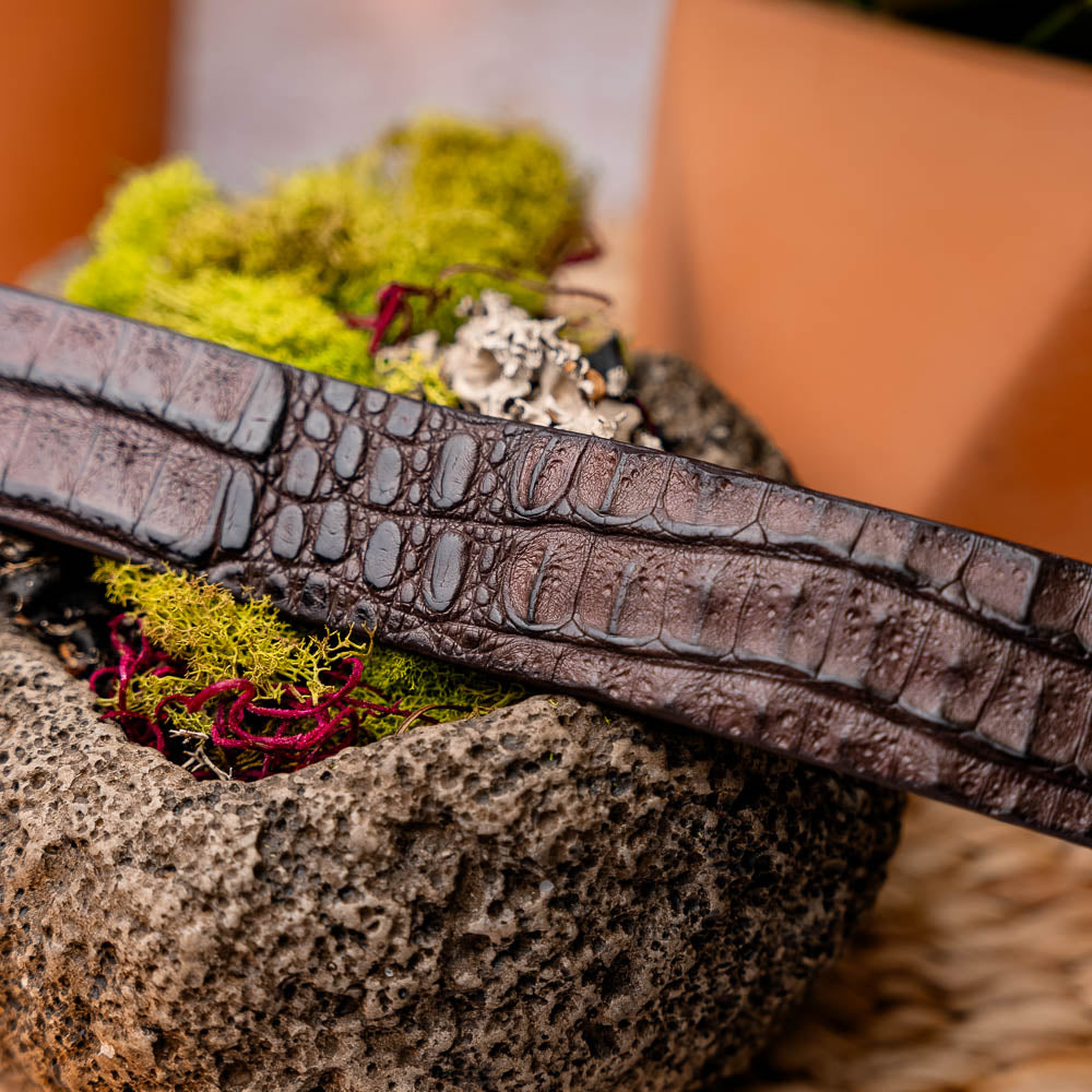 The Riccardo Belt Brown by Marco Di Milano is a dark brown exotic leather belt with a silver buckle and an engraved logo, crafted to mimic genuine caiman belly.