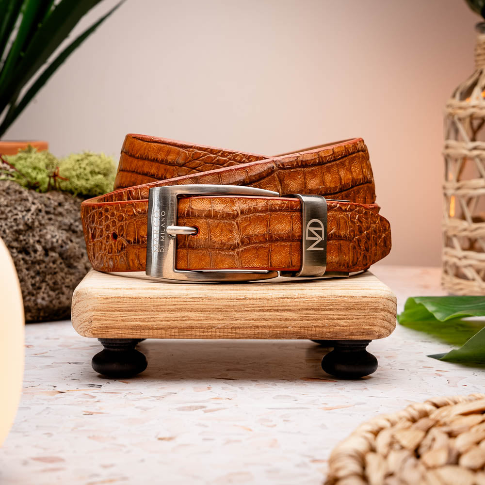 The Riccardo Belt Cognac by Marco Di Milano is a luxury exotic belt made from genuine caiman belly leather with a crocodile pattern and features a silver buckle engraved with the brand’s logo and signature.