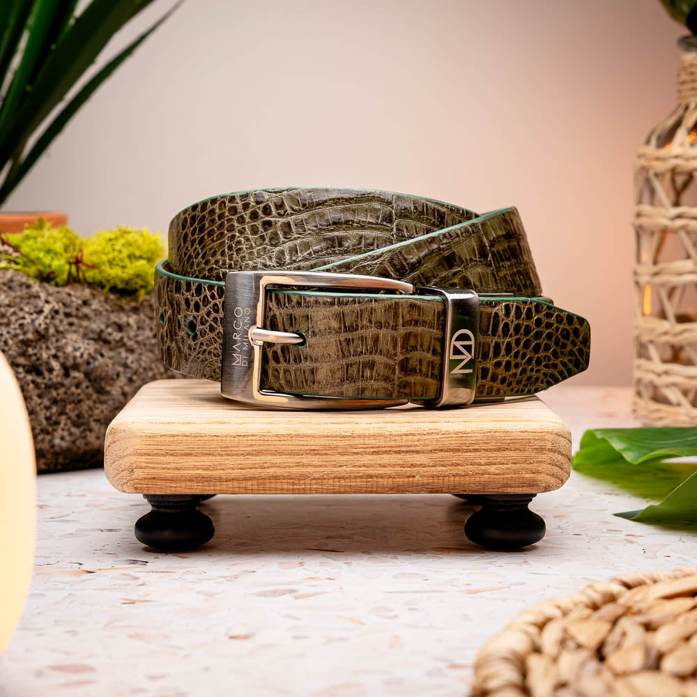 The Riccardo Belt Rustic Olive by Marco Di Milano features rustic olive caiman fuscus leather with a crocodile pattern, finished with a silver buckle engraved with the brand logo and coiled neatly.