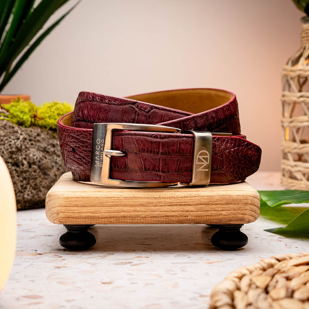The Riccardo Belt Wine by Marco Di Milano is a maroon caiman belly leather belt with a crocodile pattern and a silver buckle engraved with the brand name and logo—an elegant choice for refined style.