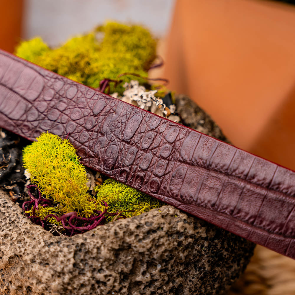 The Riccardo Belt Wine by Marco Di Milano is a maroon caiman belly leather belt with a crocodile pattern and a silver buckle engraved with the brand name and logo—an elegant choice for refined style.