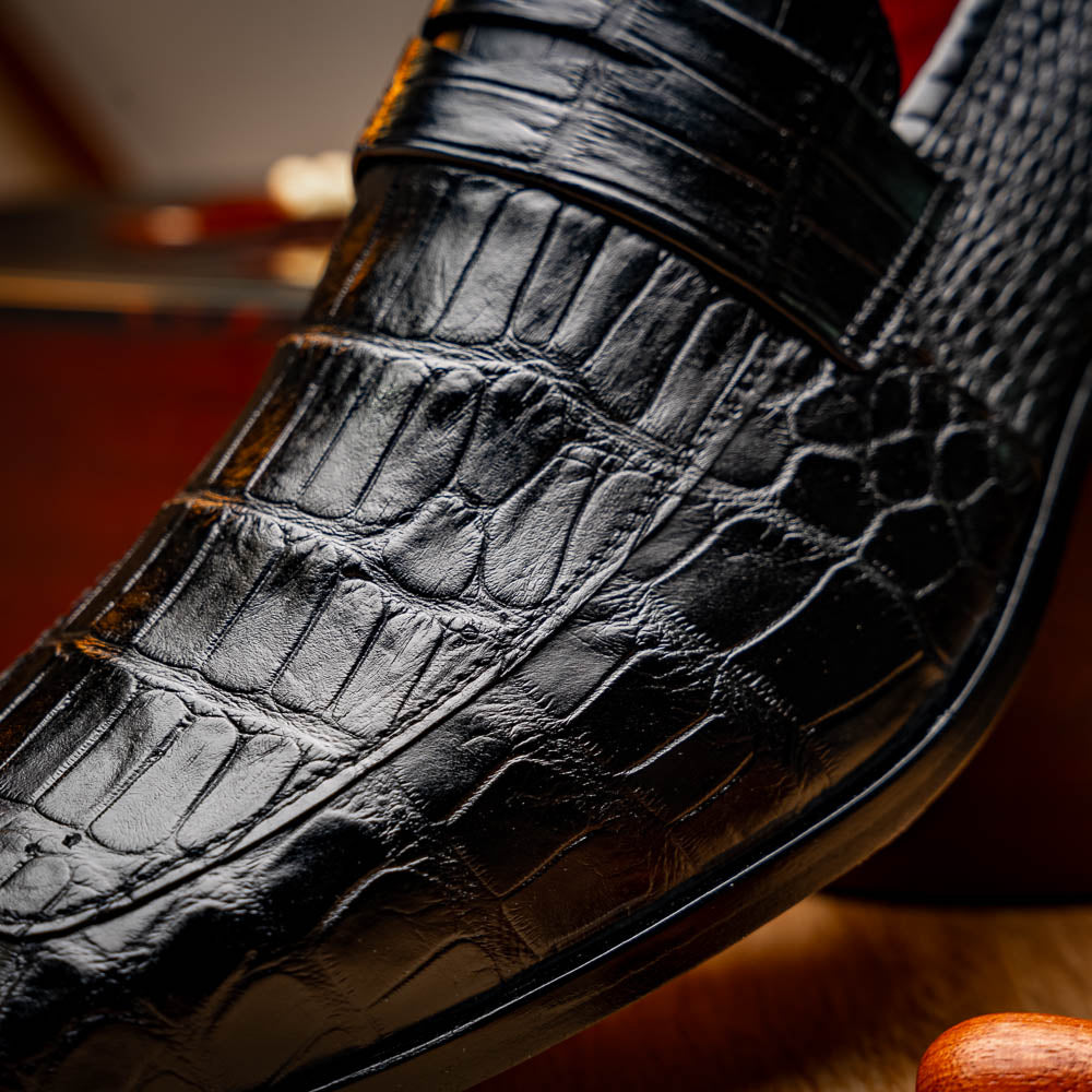 The Matteo Black by Marco Di Milano is a pair of black genuine alligator slip-on shoes with a polished brown leather sole, embossed branding, and hand-painted finishes, shown against a white background.