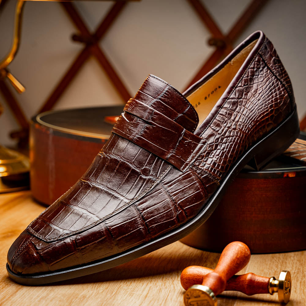 The Matteo Brown by Marco Di Milano is a pair of brown luxury loafers with a crocodile pattern; one shoe lies flat while the other is propped to showcase the sole and elegant hand-painted finishes.