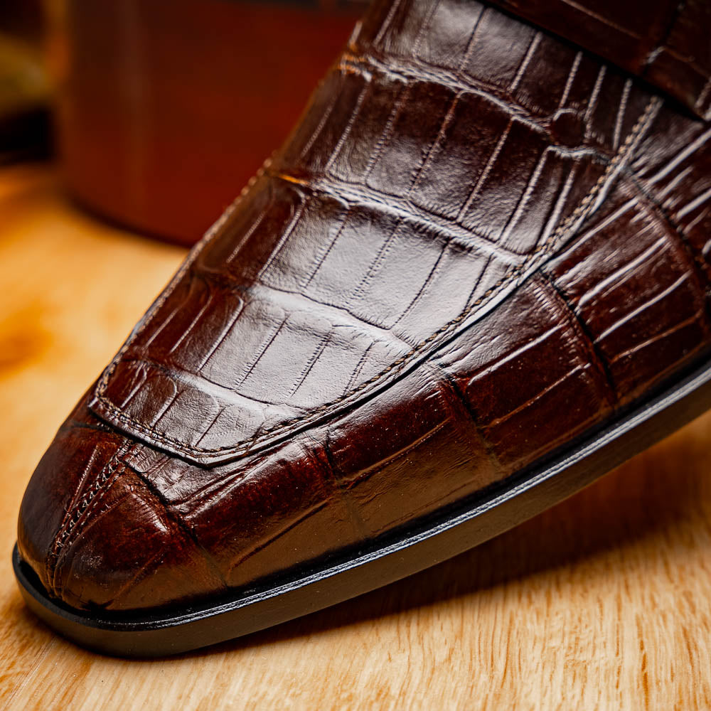 The Matteo Brown by Marco Di Milano is a pair of brown luxury loafers with a crocodile pattern; one shoe lies flat while the other is propped to showcase the sole and elegant hand-painted finishes.
