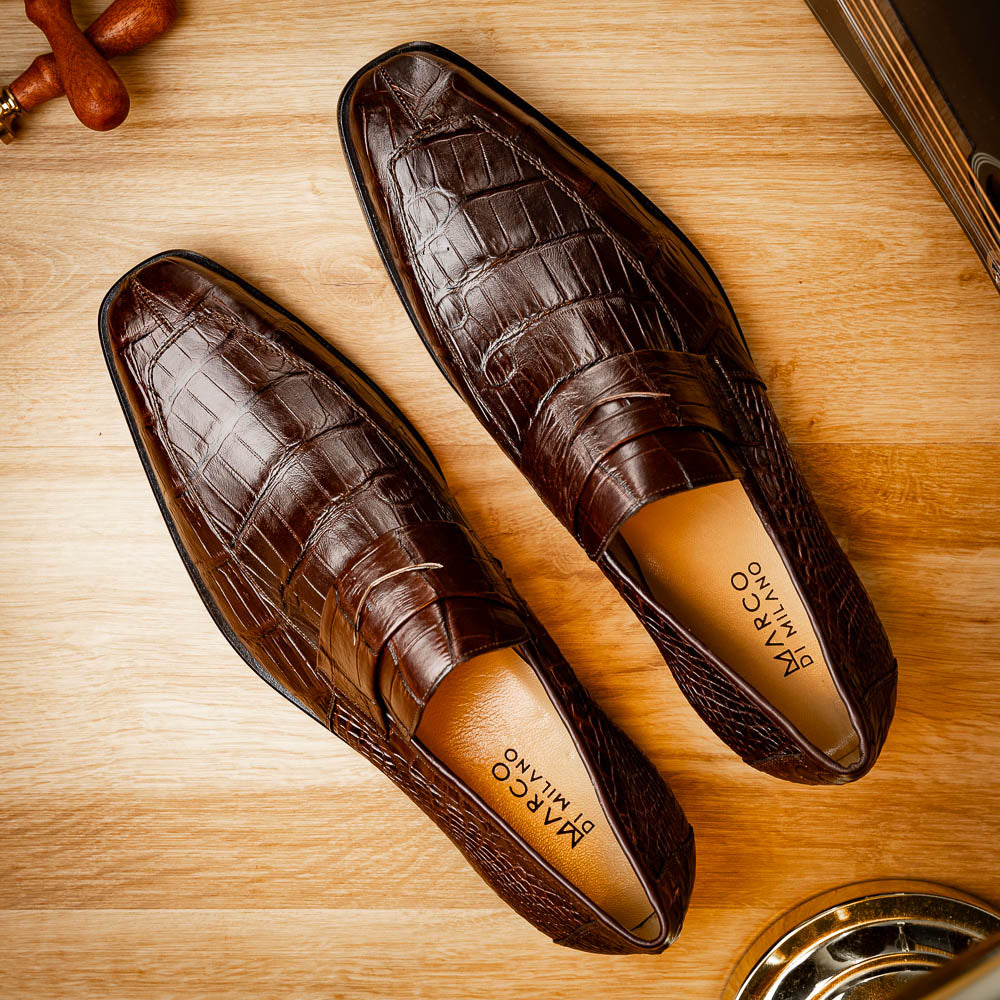 The Matteo Brown by Marco Di Milano is a pair of brown luxury loafers with a crocodile pattern; one shoe lies flat while the other is propped to showcase the sole and elegant hand-painted finishes.