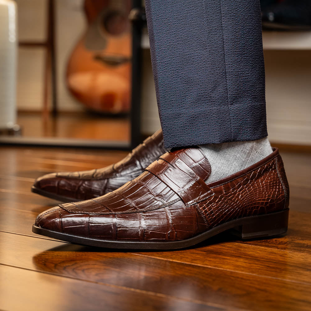 The Matteo Brown by Marco Di Milano is a pair of brown luxury loafers with a crocodile pattern; one shoe lies flat while the other is propped to showcase the sole and elegant hand-painted finishes.