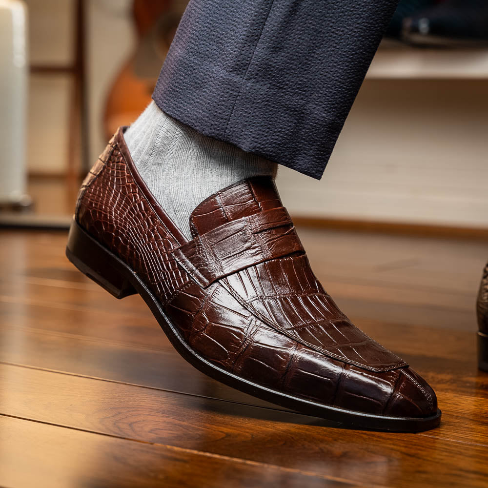 The Matteo Brown by Marco Di Milano is a pair of brown luxury loafers with a crocodile pattern; one shoe lies flat while the other is propped to showcase the sole and elegant hand-painted finishes.