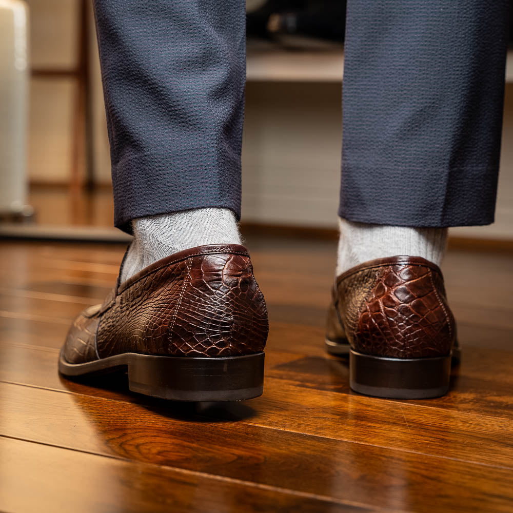 The Matteo Brown by Marco Di Milano is a pair of brown luxury loafers with a crocodile pattern; one shoe lies flat while the other is propped to showcase the sole and elegant hand-painted finishes.