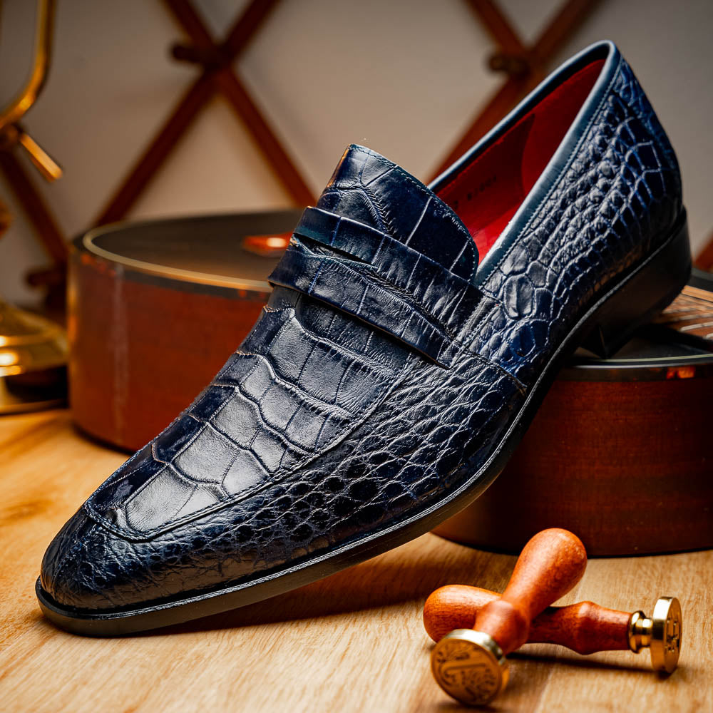 A pair of Matteo Navy loafers by Marco Di Milano, crafted in crocodile-pattern navy leather with a penny strap, red interior lining, and black soles; one shoe props against the other to reveal the sole.