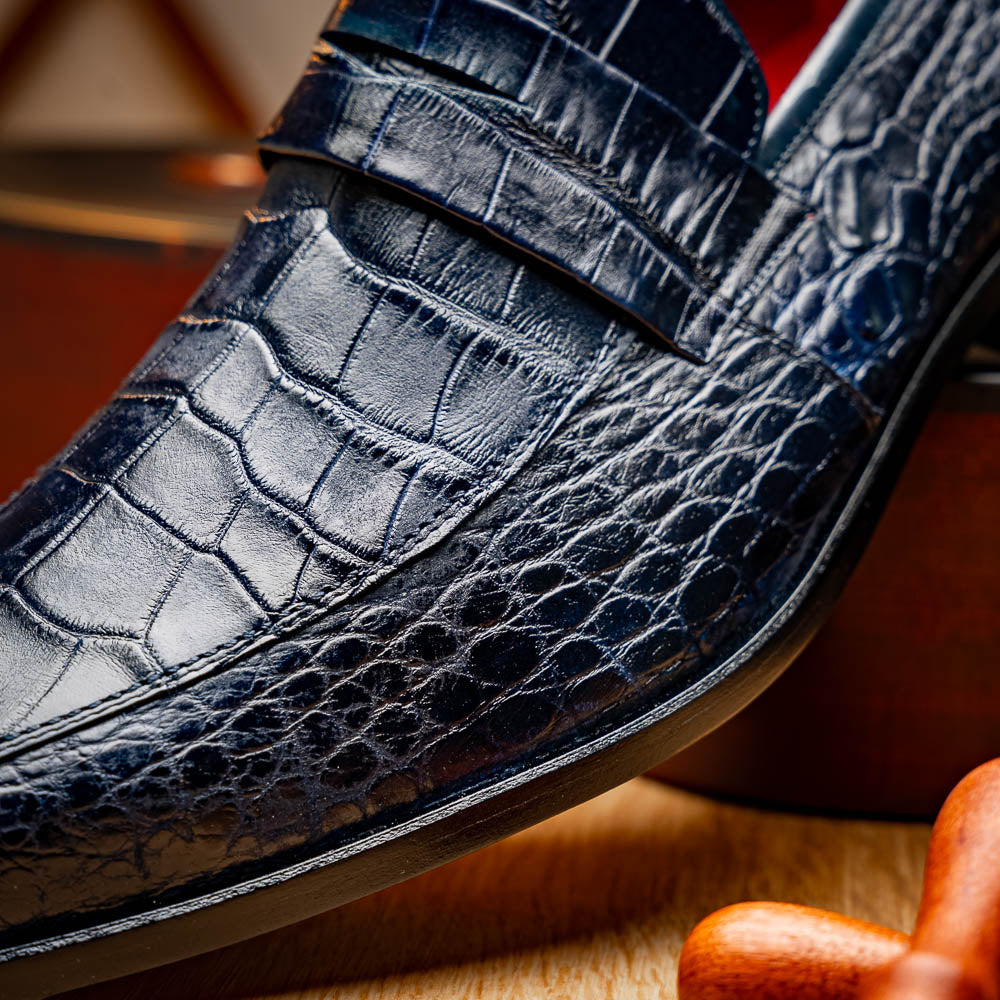 A pair of Matteo Navy loafers by Marco Di Milano, crafted in crocodile-pattern navy leather with a penny strap, red interior lining, and black soles; one shoe props against the other to reveal the sole.