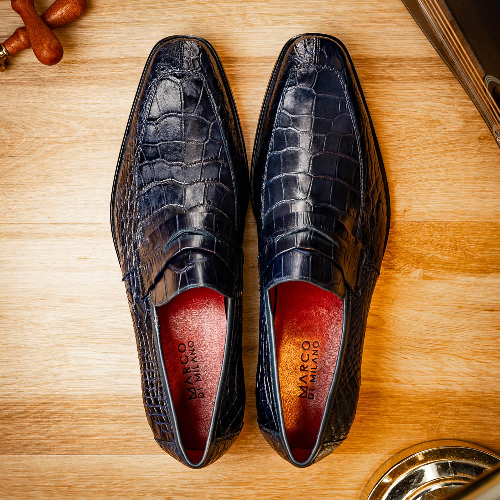 A pair of Matteo Navy loafers by Marco Di Milano, crafted in crocodile-pattern navy leather with a penny strap, red interior lining, and black soles; one shoe props against the other to reveal the sole.