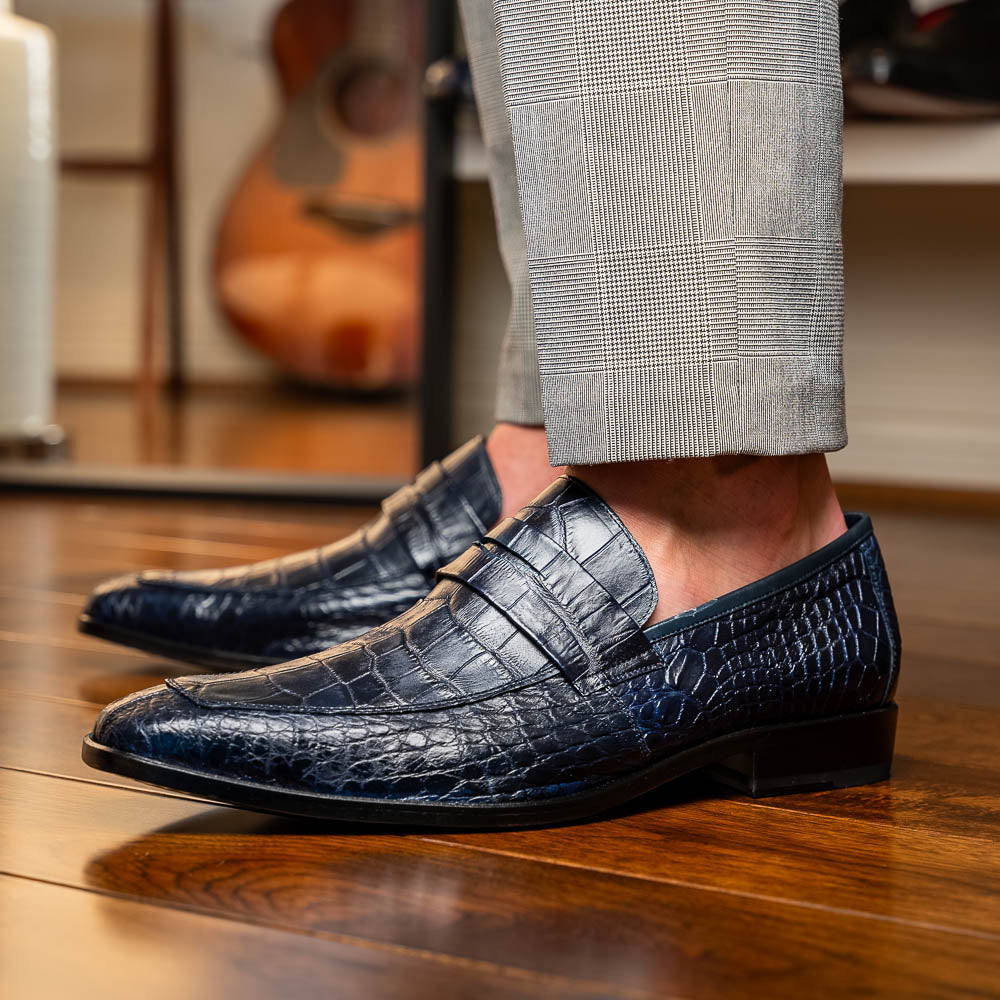 A pair of Matteo Navy loafers by Marco Di Milano, crafted in crocodile-pattern navy leather with a penny strap, red interior lining, and black soles; one shoe props against the other to reveal the sole.