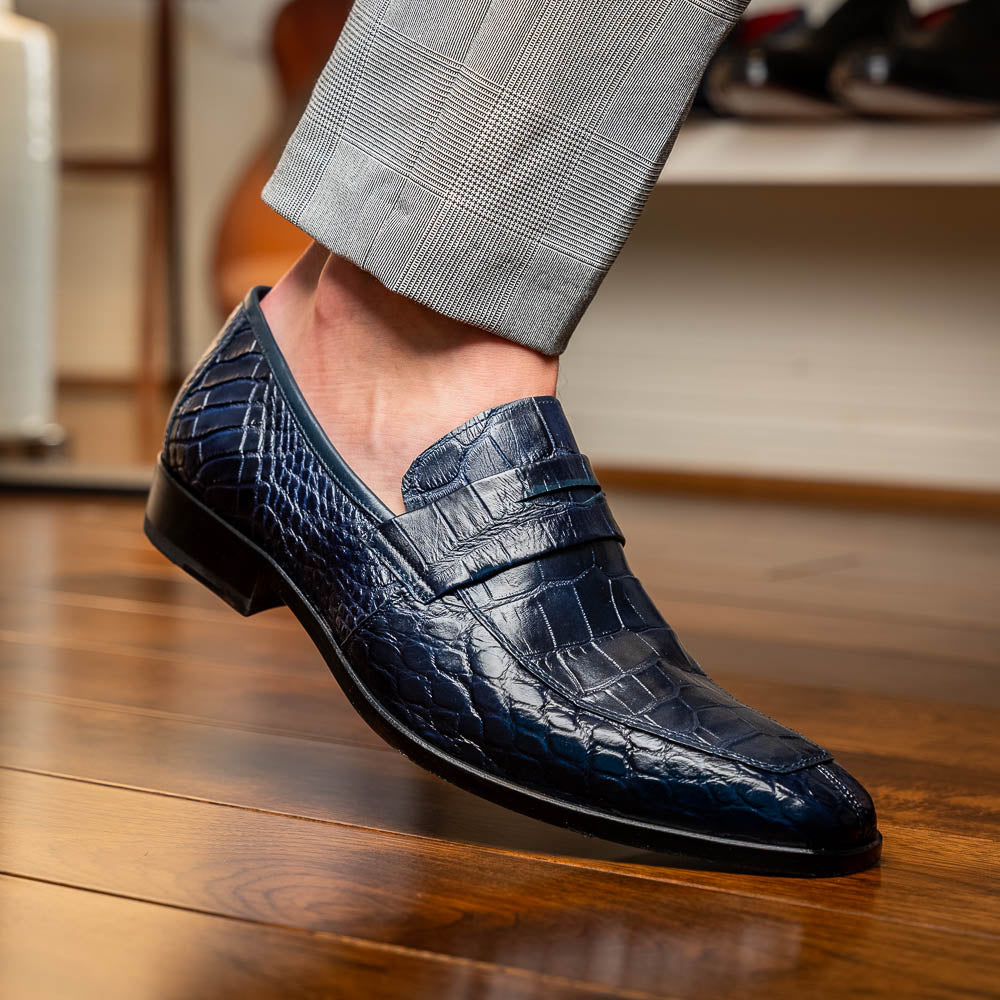 A pair of Matteo Navy loafers by Marco Di Milano, crafted in crocodile-pattern navy leather with a penny strap, red interior lining, and black soles; one shoe props against the other to reveal the sole.
