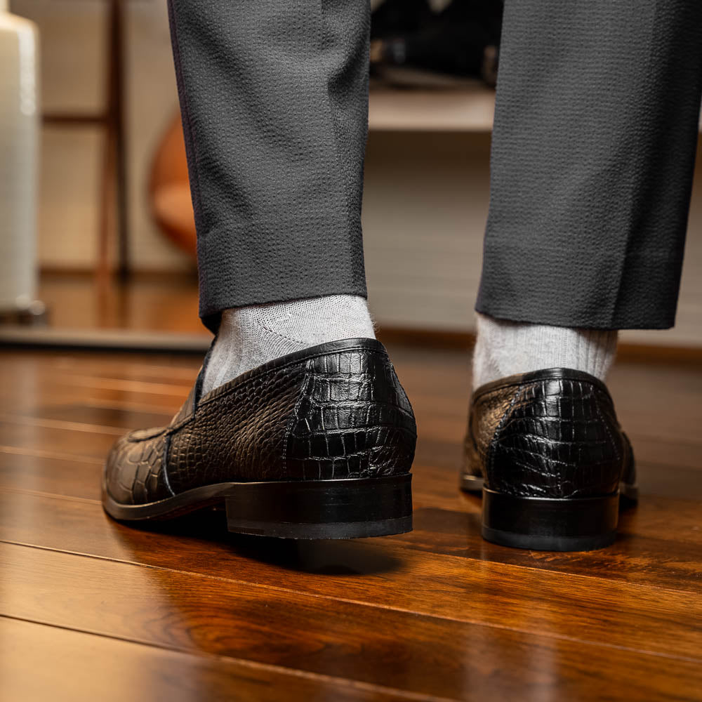 The Matteo Black by Marco Di Milano is a pair of black genuine alligator slip-on shoes with a polished brown leather sole, embossed branding, and hand-painted finishes, shown against a white background.