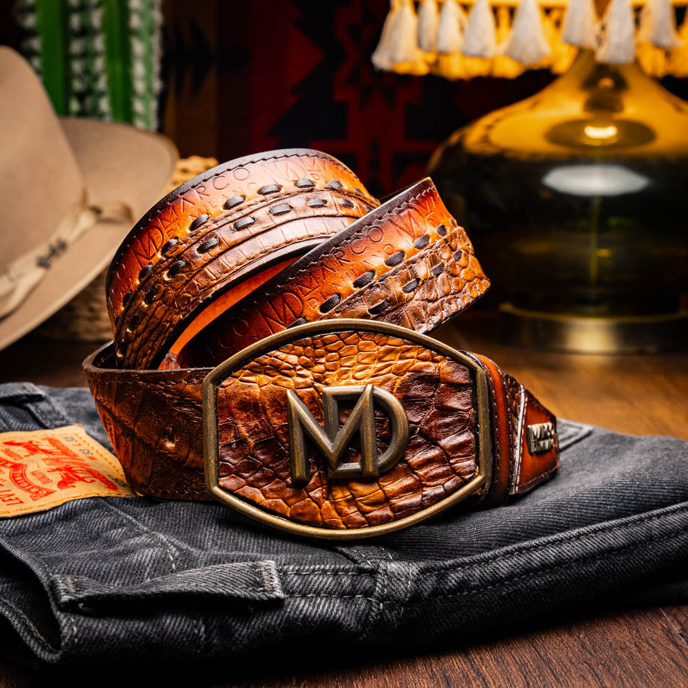 The Montana Belt Aged Maple by Marco Di Milano, featuring stitched details and a large gold MD monogram buckle with embossed branding, is coiled against a white background.