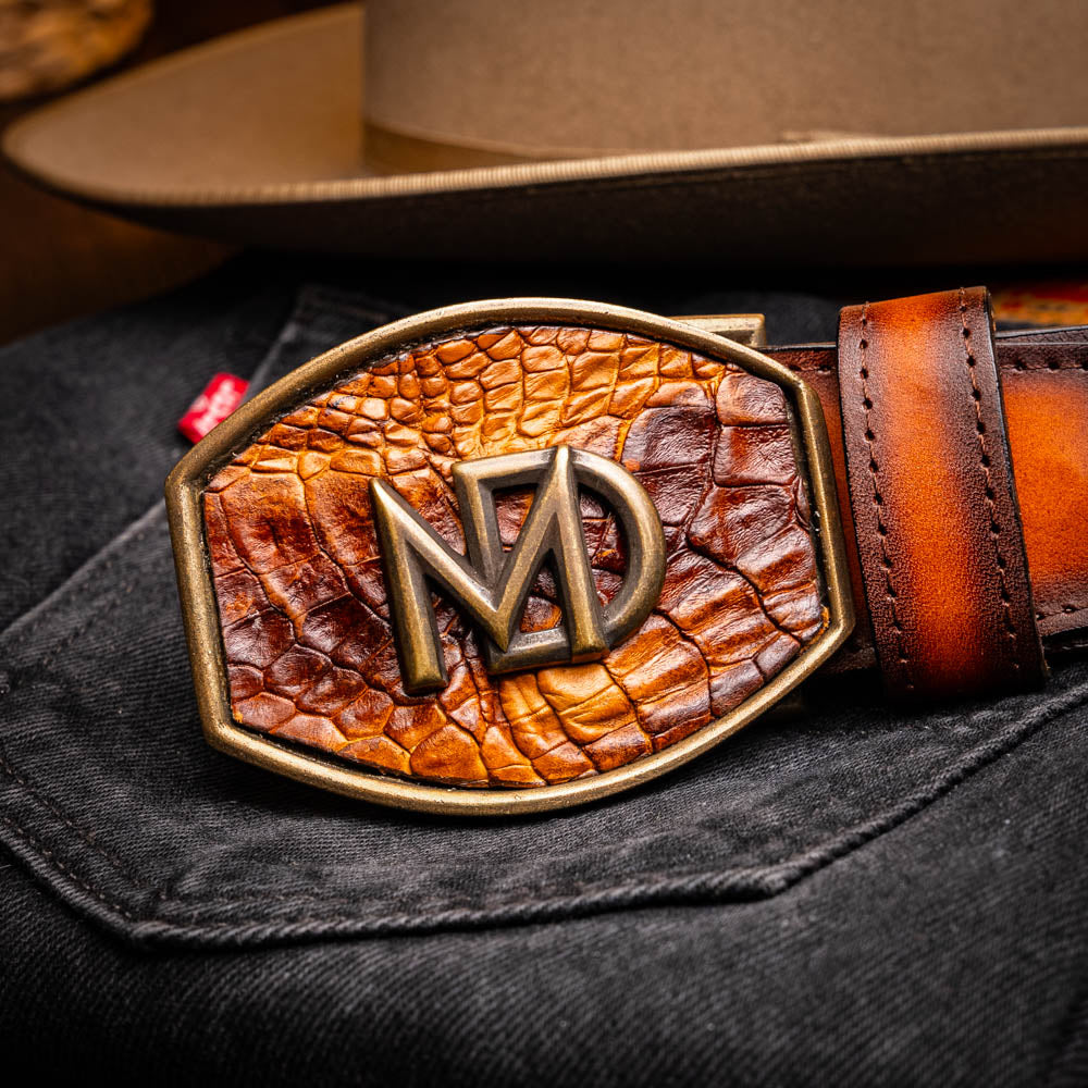 The Montana Belt Aged Maple by Marco Di Milano, featuring stitched details and a large gold MD monogram buckle with embossed branding, is coiled against a white background.