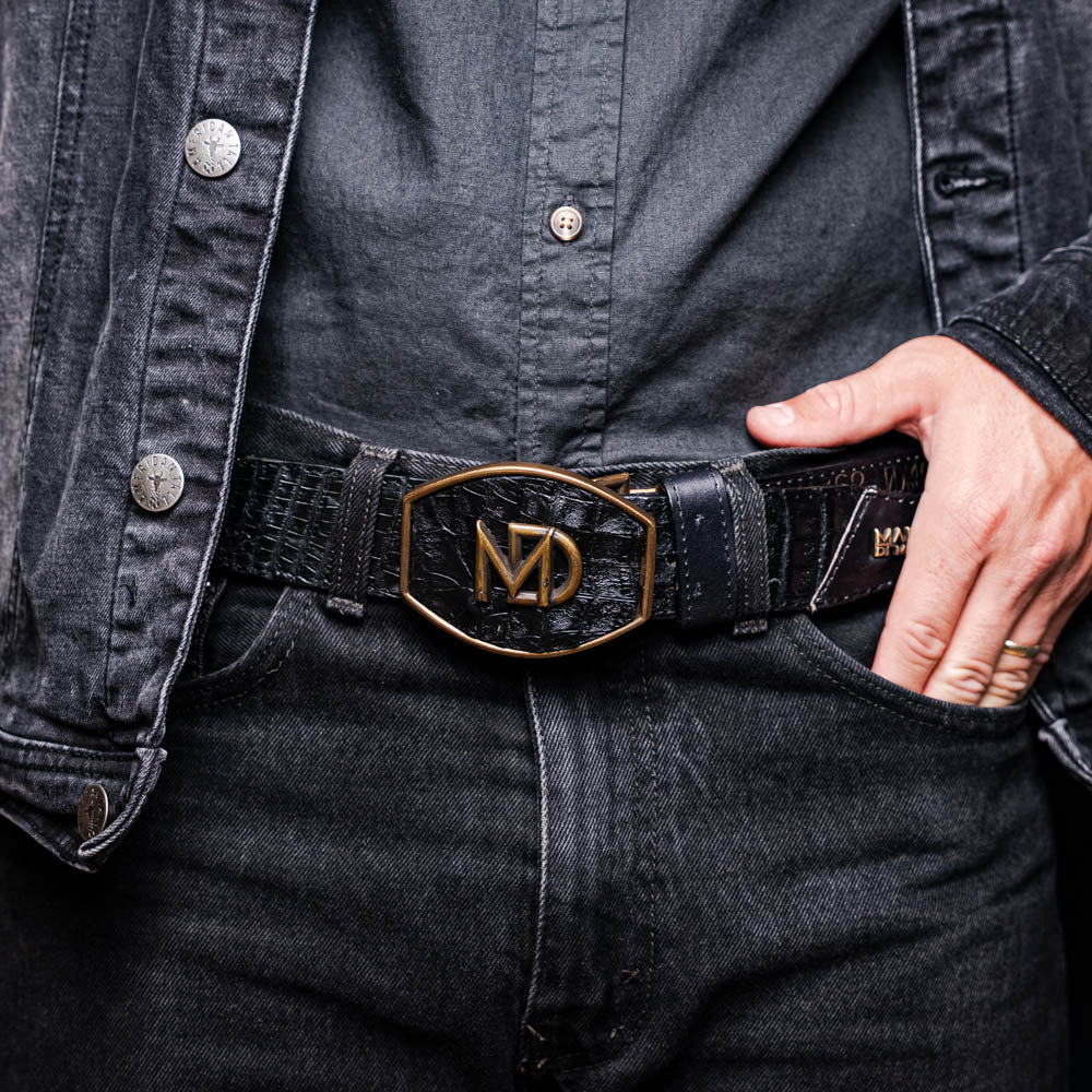 The Marco Di Milano Montana Belt Black features handcrafted black textured leather and a large gold MD monogram buckle, elegantly coiled against a white background.