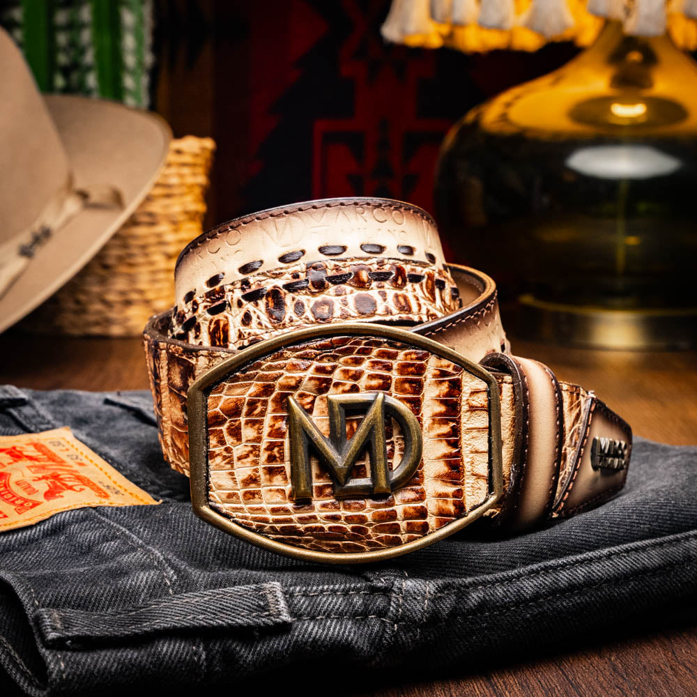 The Montana Belt Stone Orix by Marco Di Milano is a brown handcrafted leather belt with a caiman crocodile pattern and a bold gold MD monogram buckle, elegantly coiled on a white background.