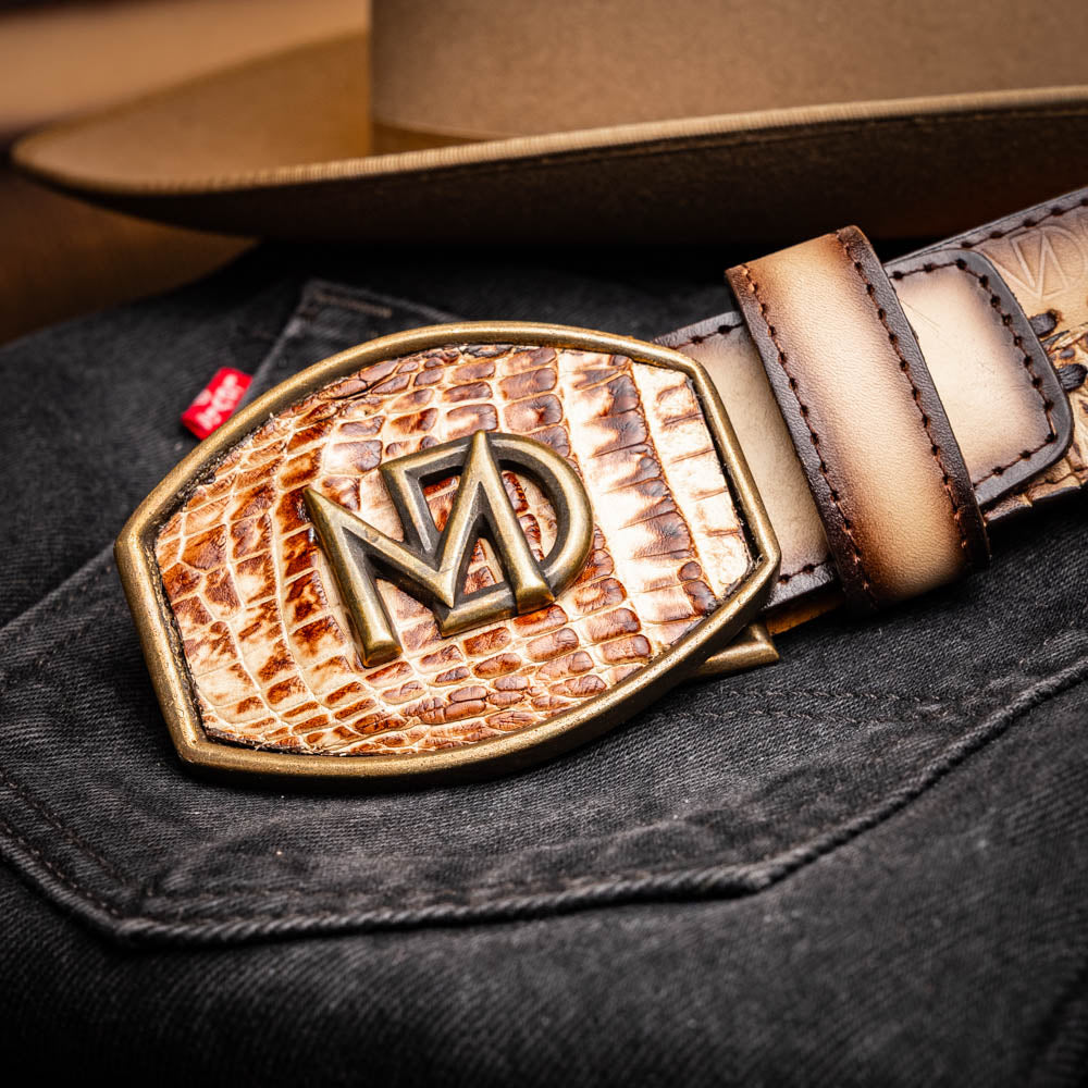 The Montana Belt Stone Orix by Marco Di Milano is a brown handcrafted leather belt with a caiman crocodile pattern and a bold gold MD monogram buckle, elegantly coiled on a white background.